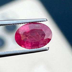 Op de afbeelding: Een levendige, ovaal geslepen robijn, vastgehouden door een zilveren pincet. De robijn vertoont een dieprode kleur met zichtbare interne facetten. De achtergrond is een vage mix van wit en blauw.