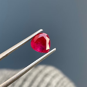 Könnte beinhalten: Ein leuchtend roter birnenförmiger Edelstein, der von einer Pinzette gehalten wird. Der Edelstein ist ein Rubin, ein Edelstein, der für seine tiefrote Farbe und seinen Glanz bekannt ist.