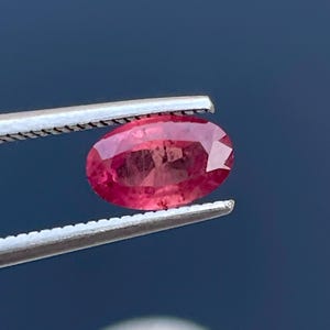 Puede incluir: Una gema roja facetada, de forma ovalada, es sostenida por unas pinzas plateadas. La gema tiene un color intenso y rico y un brillo brillante, que refleja la luz. El fondo es de color azul oscuro sólido.
