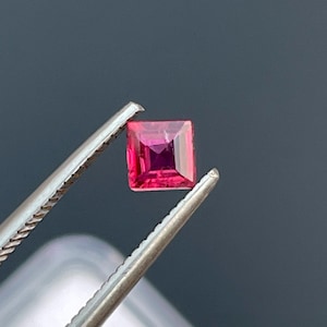 May include: A square-cut, vibrant red gemstone held by silver tweezers. The gemstone has a deep, rich color with facets that catch the light. The tweezers are in sharp focus, highlighting the gemstone's clarity and brilliance. The background is a dark gray.
