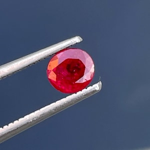 Op de afbeelding: Een helderrode edelsteen, waarschijnlijk een robijn, wordt vastgehouden in een pincet. De edelsteen is rond en heeft een facetgeslepen vorm.