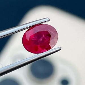 Puede incluir: Un vibrante rubí de corte ovalado sostenido por unas pinzas plateadas. El rubí muestra un tono rojo intenso con reflejos internos, lo que sugiere claridad y brillo. Las pinzas están enfocadas, resaltando los detalles de la gema.