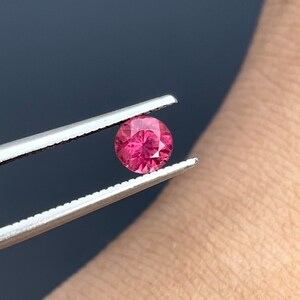 May include: A round, pink gemstone held by tweezers. The gemstone is faceted and has a brilliant shine.