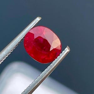 May include: A vibrant red oval-shaped gemstone held by tweezers. The gemstone is a ruby with a smooth, polished surface.