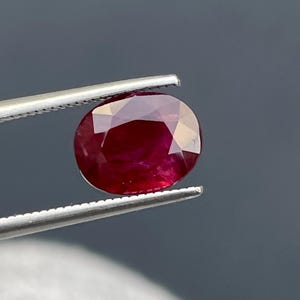 Puede incluir: Una gema de rubí facetada, de forma ovalada, sujeta con pinzas plateadas. El rubí es de un color rojo intenso y rico, con una superficie pulida que refleja la luz. El fondo es de un gris oscuro sólido.