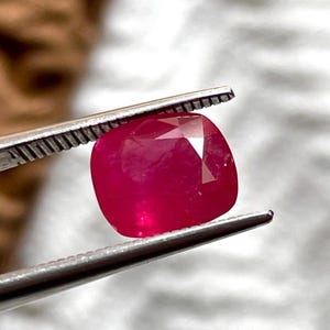 Puede incluir: Una gema de rubí de corte cojín con un tono rojo intenso y una superficie multifacetada, sostenida de forma segura entre las puntas metálicas dentadas de unas pinzas plateadas, mostrando su claridad translúcida y facetas geométricas.