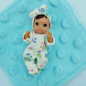 May include: A baby doll wearing a white onesie and matching headband, both adorned with a floral pattern in pink and blue. The doll is on a light blue, textured blanket.