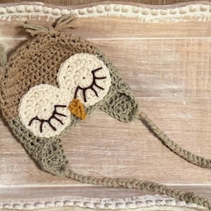 May include: A handmade crochet owl hat in shades of brown, beige, and green. The hat features large, round eyes with eyelashes and a small, yellow beak. The hat has ear flaps and long ties for securing it. A charming and whimsical accessory.