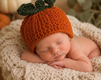 Pode incluir: Um gorro de crochê laranja feito à mão em forma de abóbora, com um caule e uma folha verde-escura no topo. O gorro é usado por um bebé a dormir num cesto de crochê de cor creme com textura. O texto ShopLenaElisabeth.etsy.com é visível.