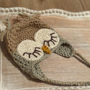 May include: A handmade crochet owl hat in shades of brown, beige, and gray. The hat features large, round eyes with eyelashes and a small yellow beak. The hat has ear flaps and ties. The hat is designed for a baby or toddler.