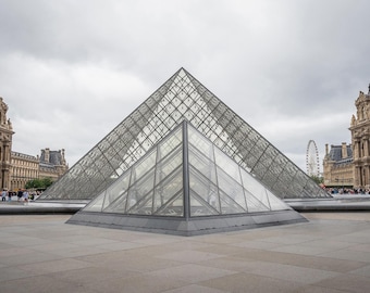 May include: A wide shot of the Louvre Museum in Paris, France, featuring the iconic glass pyramid in the center. The pyramid is surrounded by a large plaza and flanked by the historic museum buildings under a cloudy sky.
