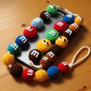 May include: A colorful phone strap made of crocheted M&M candies. The strap is attached to a smartphone with a gray case. The strap has a brown, red, yellow, green, blue, and white crocheted candy beads.