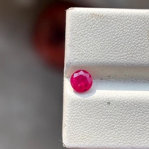 Può includere: Una pietra preziosa di rubino rotonda e sfaccettata di colore rosso intenso. La pietra preziosa è posizionata su uno sfondo bianco.