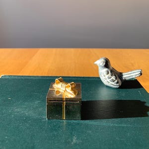 May include: A small gold gift box with a gold bow on top. The box is sitting on a green surface.