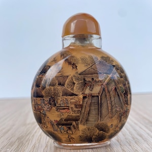 Könnte beinhalten: Eine klare Schnupftabakflasche aus Glas mit runder Form und einem braunen Verschluss. Die Flasche ist mit einer detaillierten, sepiafarbenen Landschaftsszene mit Gebäuden, Bäumen und Figuren verziert. Die Flasche steht auf einer Holzoberfläche.