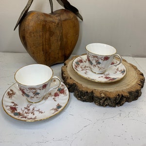 May include: Two white teacups and saucers with a floral pattern in shades of brown, pink, and blue. The cups and saucers have a gold trim. The cups and saucers are sitting on a wooden slice.