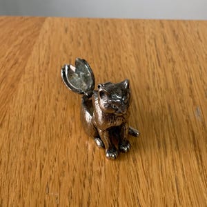 May include: A silver-colored cat figurine with an open compartment on its back. The cat is in a seated position with its tail extended. The figurine is on a wooden surface.