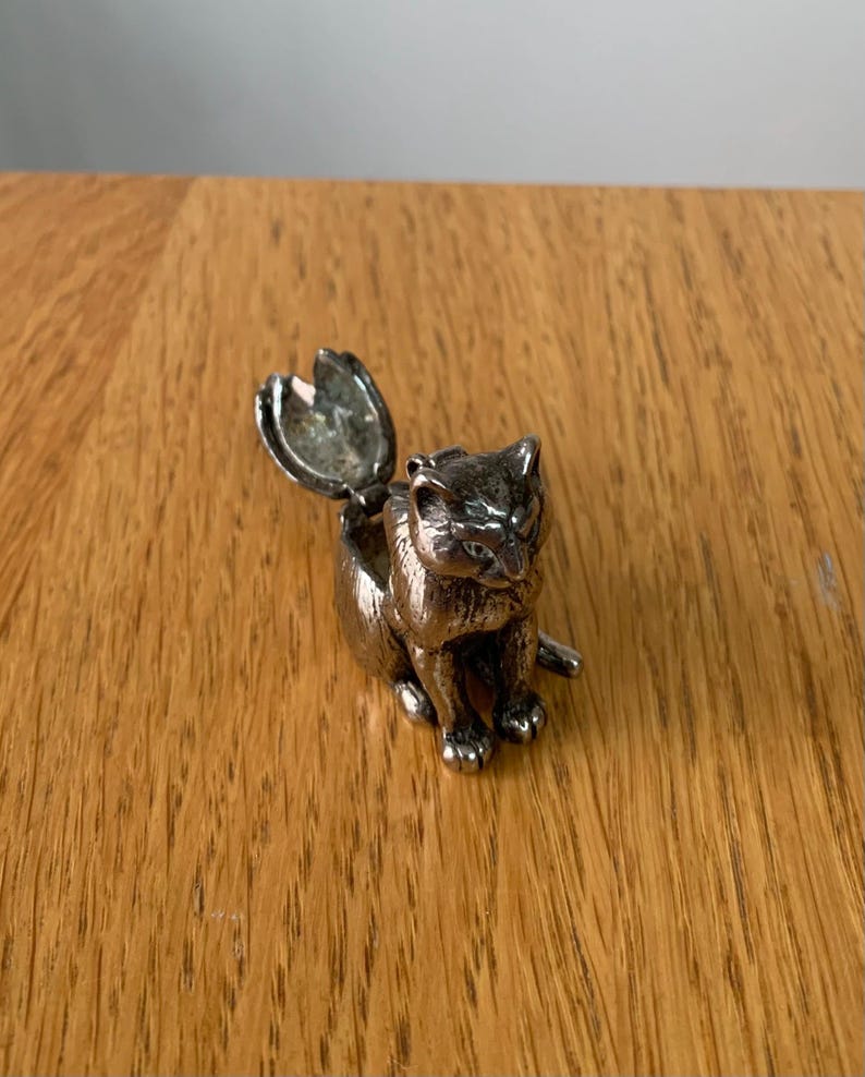 May include: A silver-colored cat figurine with an open compartment on its back. The cat is in a seated position with its tail extended. The figurine is on a wooden surface.
