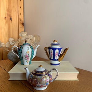 Könnte beinhalten: Drei Porzellan-Teekannen mit aufwendigen floralen und geometrischen Mustern. Die Teekannen sind weiß mit goldenen Akzenten und haben verschiedene Farben, darunter Blau, Grün und Rosa.