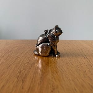 May include: A small, silver-colored cat figurine with a detailed textured surface. The cat is sitting upright with its head turned to the side. The figurine is on a wooden surface.
