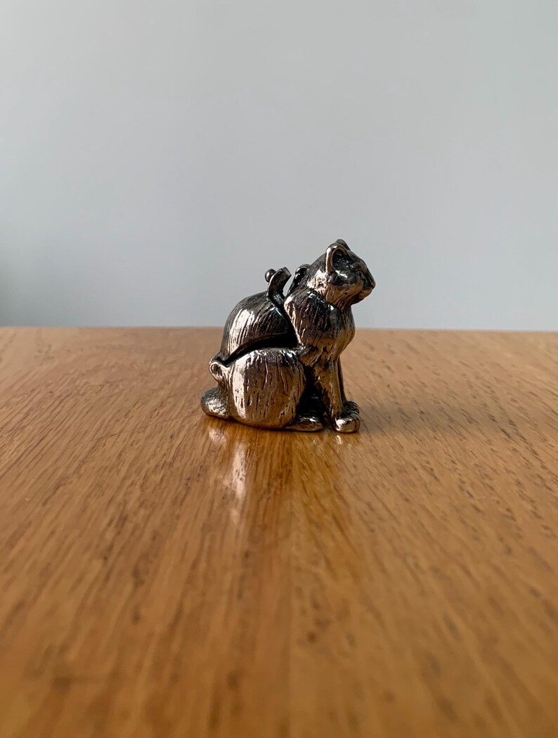 May include: A small, silver-colored cat figurine with a detailed textured surface. The cat is sitting upright with its head turned to the side. The figurine is on a wooden surface.