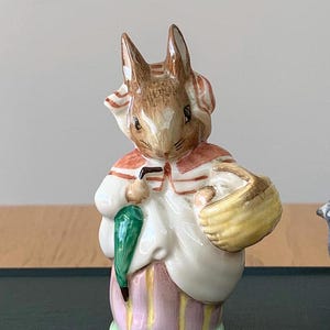May include: A porcelain figurine of a rabbit wearing a red and white striped coat, holding a green umbrella and a yellow basket. The rabbit has brown fur and a white face.