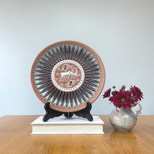 May include: A decorative plate with a brown and white antelope design, displayed on a black stand. The plate has a sunburst pattern with brown and gray accents. A small vase with dark red flowers sits nearby.