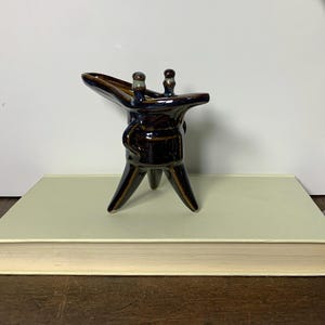 May include: A small, three-legged ceramic vessel with a brown and blue glaze. The vessel has a spout and two small handles.