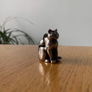 May include: A silver-colored cat figurine in a seated position. The cat has detailed features, including visible ears, eyes, and paws. The figurine is made of a metallic material and sits on a wooden surface.