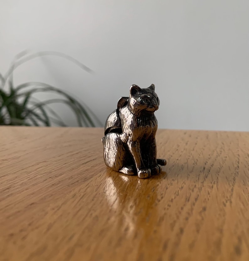May include: A silver-colored cat figurine in a seated position. The cat has detailed features, including visible ears, eyes, and paws. The figurine is made of a metallic material and sits on a wooden surface.