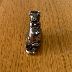 May include: A silver-colored cat-shaped trinket box. The cat is sitting upright with its tail curled. The box has a hinged opening on its belly. The cat is on a wooden surface.