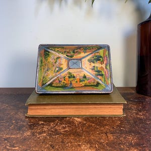 May include: Vintage tin box with a colorful image of a garden scene. The box is rectangular and has a blue, green, and yellow color scheme. The image depicts a garden with flowers, trees, and a fountain.