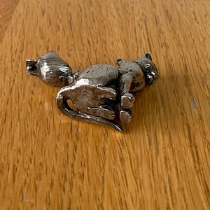 May include: A silver-colored cat figurine in a resting pose. The cat is detailed with visible fur texture and is lying on its side. The figurine is placed on a wooden surface with a natural wood grain pattern.