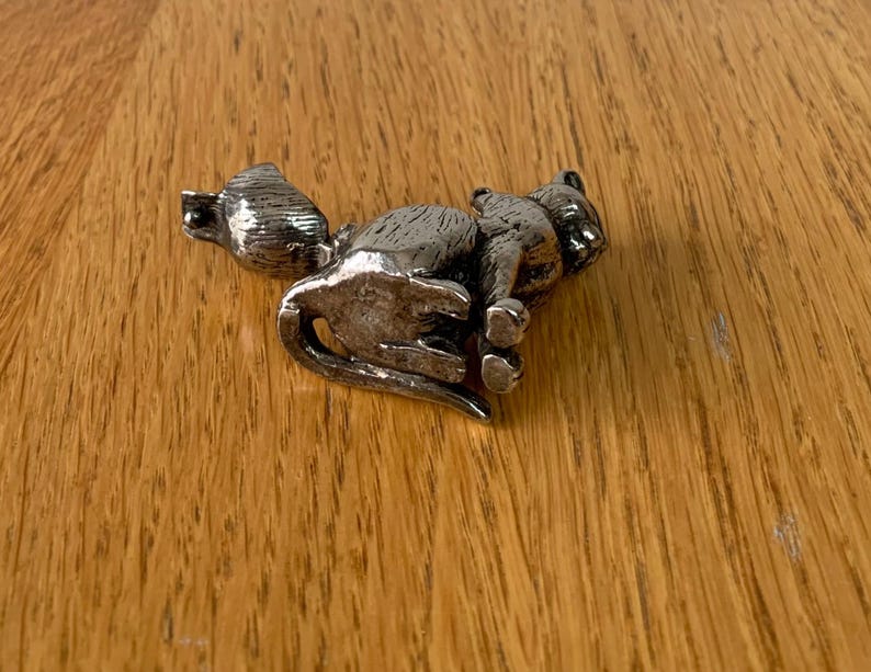May include: A silver-colored cat figurine in a resting pose. The cat is detailed with visible fur texture and is lying on its side. The figurine is placed on a wooden surface with a natural wood grain pattern.