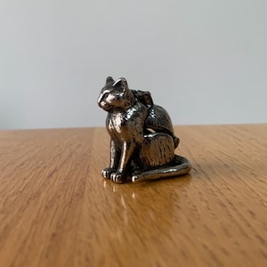 May include: A small, silver-colored cat figurine in a seated position. The cat has detailed features and is sitting on a wooden surface. The figurine is a decorative item, possibly for a shelf or desk.