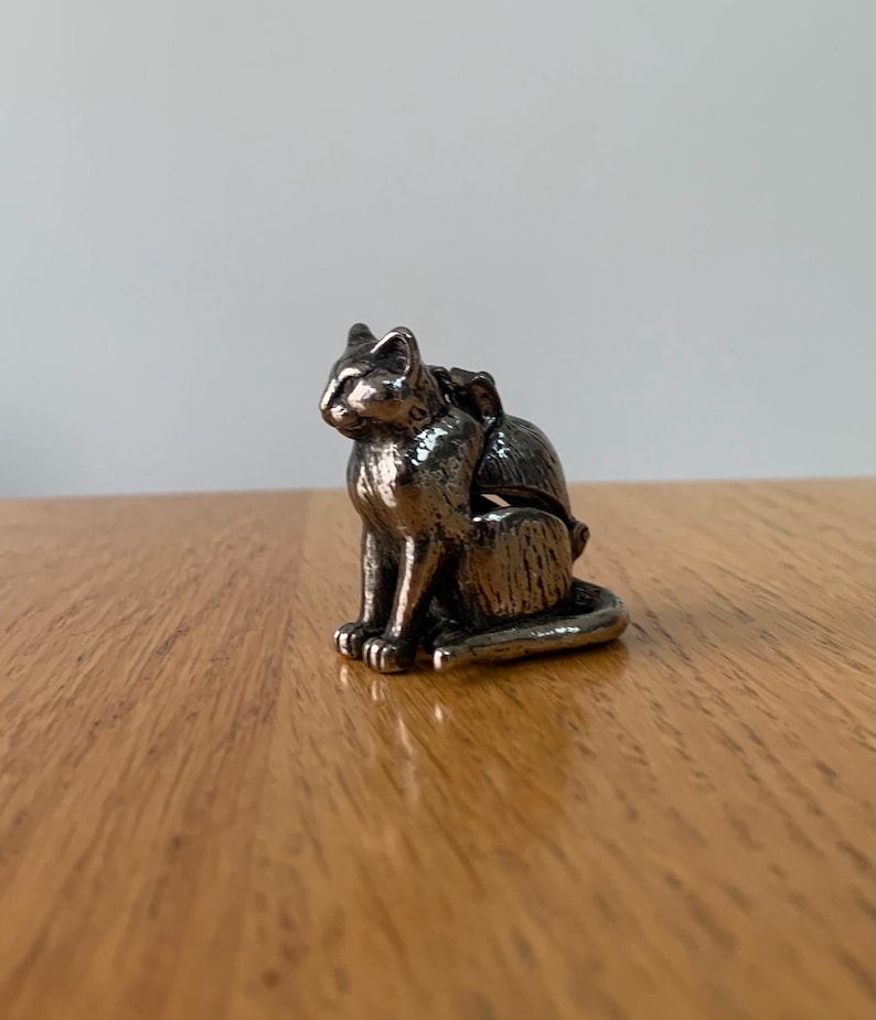 May include: A small, silver-colored cat figurine in a seated position. The cat has detailed features and is sitting on a wooden surface. The figurine is a decorative item, possibly for a shelf or desk.