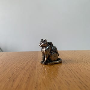 May include: A small, silver-colored cat figurine in a seated position. The cat has detailed features, including pointed ears and a curled tail. The figurine is placed on a wooden surface, with a plain white background.