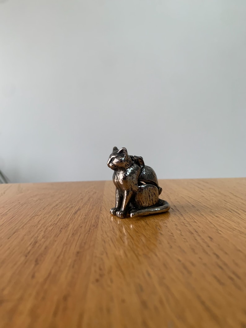 May include: A small, silver-colored cat figurine in a seated position. The cat has detailed features, including pointed ears and a curled tail. The figurine is placed on a wooden surface, with a plain white background.