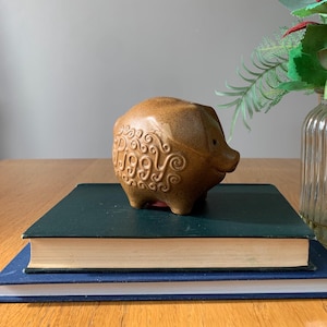May include: A brown ceramic piggy bank with the word "Piggy" inscribed on the side. The piggy bank is sitting on top of two books, one green and one blue.