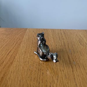 May include: A silver-colored cat-shaped trinket box with a hinged lid. The cat is in a seated position, with its tail curled. The box is made of metal and has a detailed design. The cat is on a wooden surface.