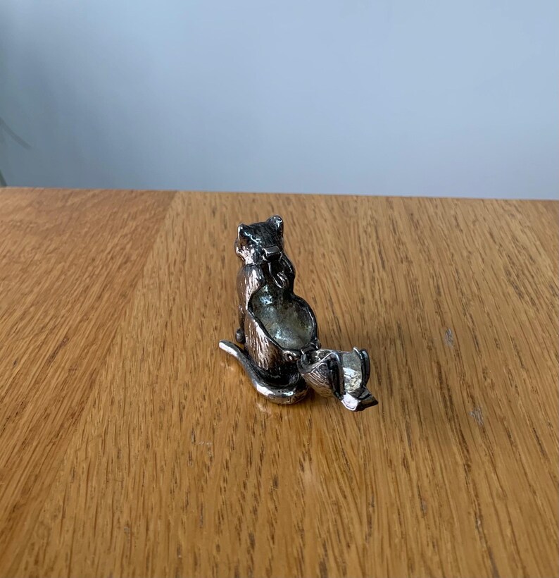 May include: A silver-colored cat-shaped trinket box with a hinged lid. The cat is in a seated position, with its tail curled. The box is made of metal and has a detailed design. The cat is on a wooden surface.