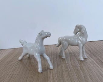 May include: Two small, white ceramic horse figurines. One horse is posed with its front legs extended, and the other stands with its head turned. The figurines are on a light brown wooden surface.