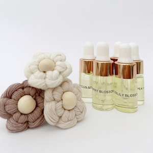 May include: Five small glass bottles with gold lids and droppers filled with a clear yellow liquid. The bottles are labelled with different scents: "Cactus", "Strawberry Blossom", "Lily Blossom", "Pea +", and "Vanilla Bean". Three small white and brown crocheted flowers with wooden centres are in the foreground.