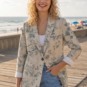 May include: Beige blazer with a floral pattern in blue and green. The blazer has a single-button closure and is worn with blue jeans. The sleeves are cuffed to reveal a white lining. The background shows a beach scene.