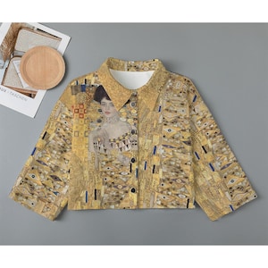 May include: A gold and yellow patterned button-up shirt with a portrait of a woman in a gold dress. The shirt has a collar and long sleeves.