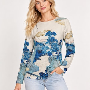 May include: A long-sleeve crew neck shirt featuring a beige base color with a detailed blue and tan landscape print. The graphic depicts stylized trees, rolling hills, and clouds in a traditional art style, accented by vertical Japanese text.