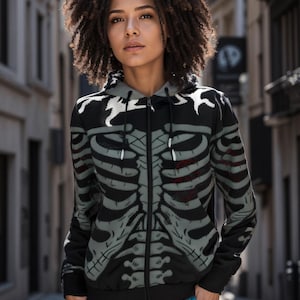 Könnte beinhalten: Schwarzer Kapuzenpullover mit Reißverschluss und einem grauen Skelett-Druck. Der Hoodie hat eine Kapuze und einen Reißverschluss. Das Model trägt eine blaue Hose.