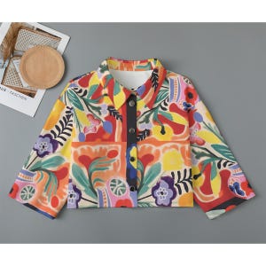 May include: A colorful button-up shirt with a floral pattern in shades of red, yellow, blue, green, and pink. The shirt has a cropped length and long sleeves.