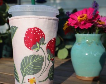Könnte beinhalten: Eine rosa-weiße Stoff-Becherhülle mit einem Erdbeer- und Blattdesign. Die Hülle befindet sich auf einem durchsichtigen Plastikbecher mit einem grünen Strohhalm.
