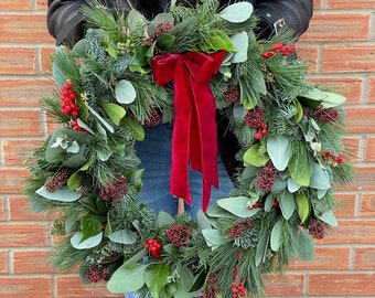 May include: A festive Christmas wreath featuring a mix of evergreen foliage, red berries, and eucalyptus leaves. A vibrant red velvet ribbon is tied in a bow at the center. The wreath is held in front of a brick wall.